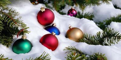 The primary subject of the image is a group of Christmas ornaments, including gold, blue, and red balls, placed on a snow-covered surface. The setting is a winter scene with snow-covered trees and a pine tree in the background. The visual style is a photo of the ornaments on the snow, capturing the festive atmosphere of the holiday season. The colors and mood of the image are predominantly cold and wintry, with the gold, blue, and red ornaments adding a touch of
